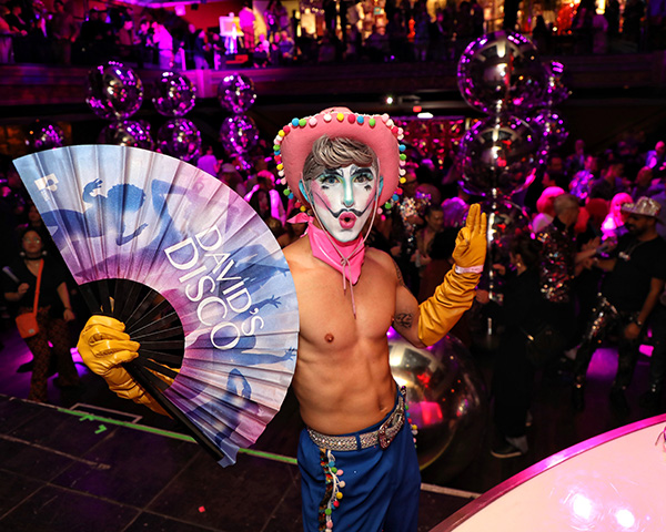 DD_DiscoCowboy shirtless, fit, young man with makeup that looks like a stylized cowboy with droopy mustache, holding a fan and wearing a pink cowboy had with colorful bobbles, under purply disco lights.