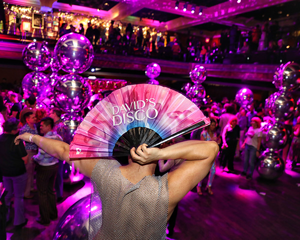 DD_Fan_600x480 person in sparkly silver shirt dancing with a fan that reads David's Disco, under purply disco lights