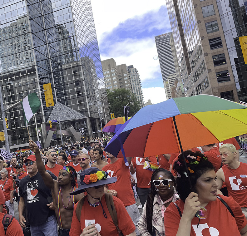 A diverse group of people marching amongst tall buildings in downtown Toronto. Most people are wearing red Casey House T-shirts and a woman in the foreground is carrying a rainbow umbrella.