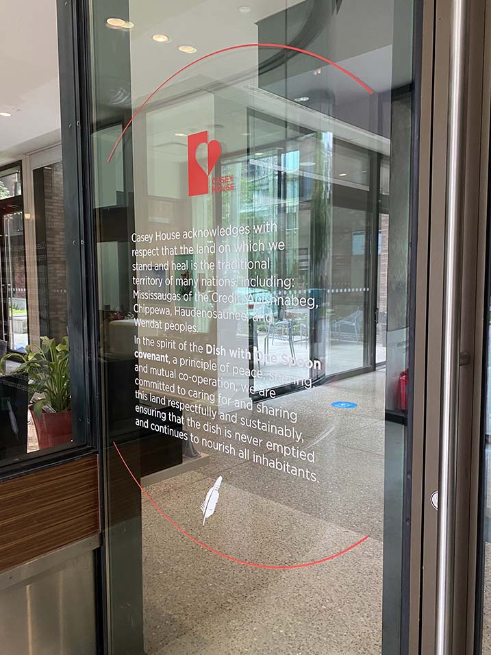 Land_Acknowledgement_IMG_4676-Edit an image of the land acknowledgement on an interior glass door. The Casey Logo is at the top followed by the acknowledgement in white with an image of a white eagle feather placed after the acknowledgment.